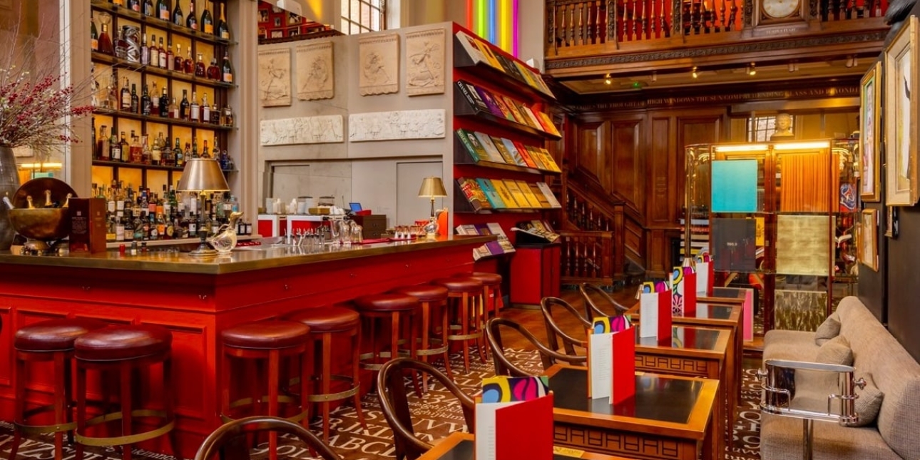 The interior of the Maison Assouline, with tables and chairs, menus, a bar with stools around it and lamps on top, and shelves full of bottles and books in the background